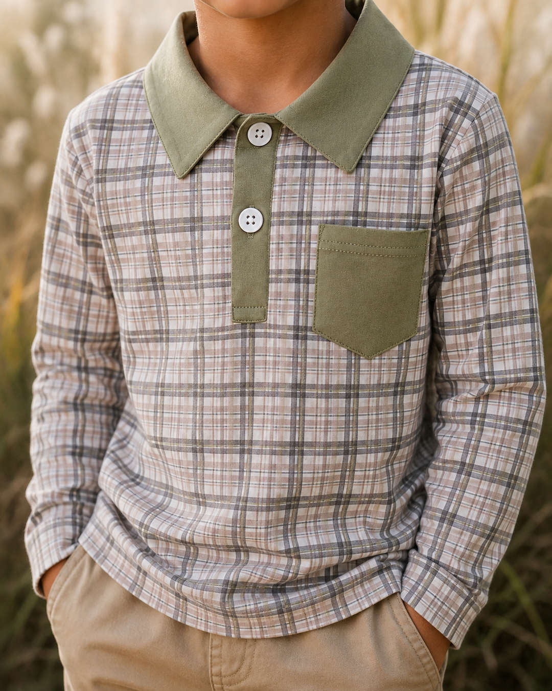 A child wearing a long-sleeve plaid button-up shirt with an olive green collar, placket, and chest pocket, paired with khaki pants, set against a soft-focus outdoor background.