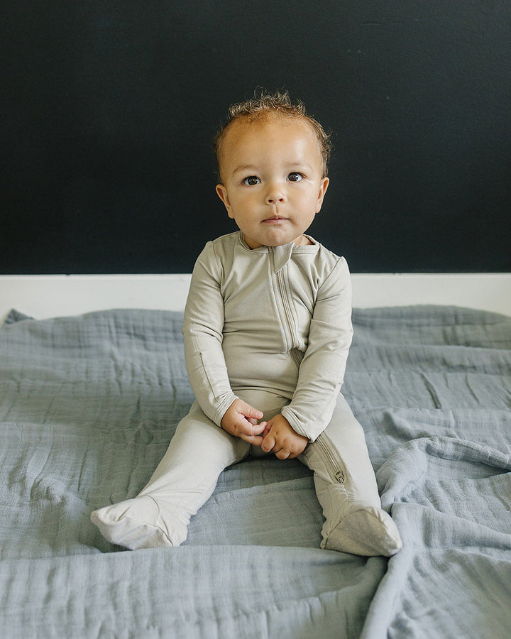 Baby sitting on a bed wearing a beige onesie against a dark background