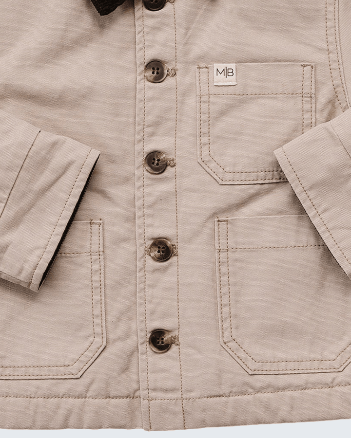 Close-up of a beige canvas kids jacket with brown buttons, featuring a chest pocket with a white &