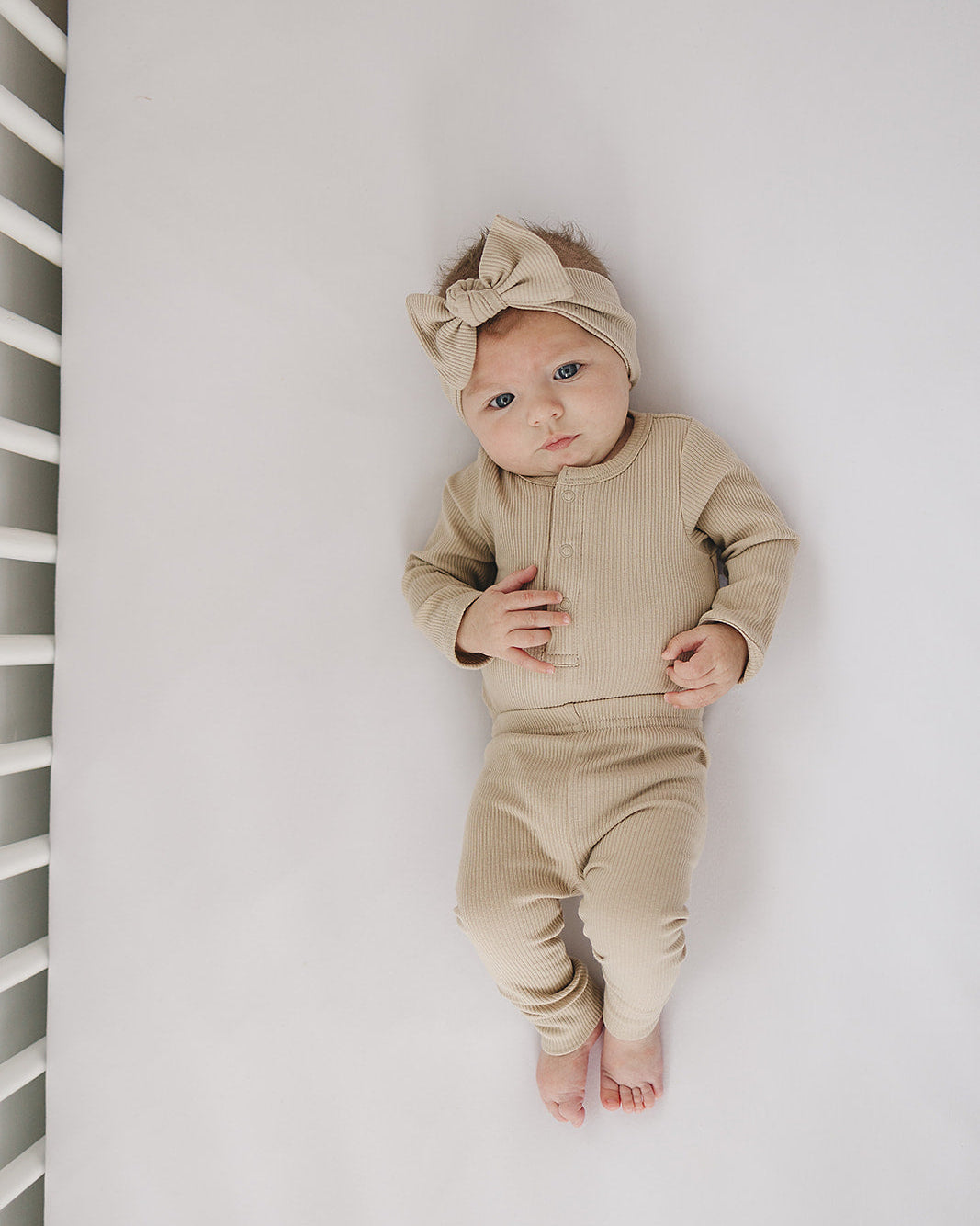 Baby in a beige outfit lying on a white surface with a white crib in the background