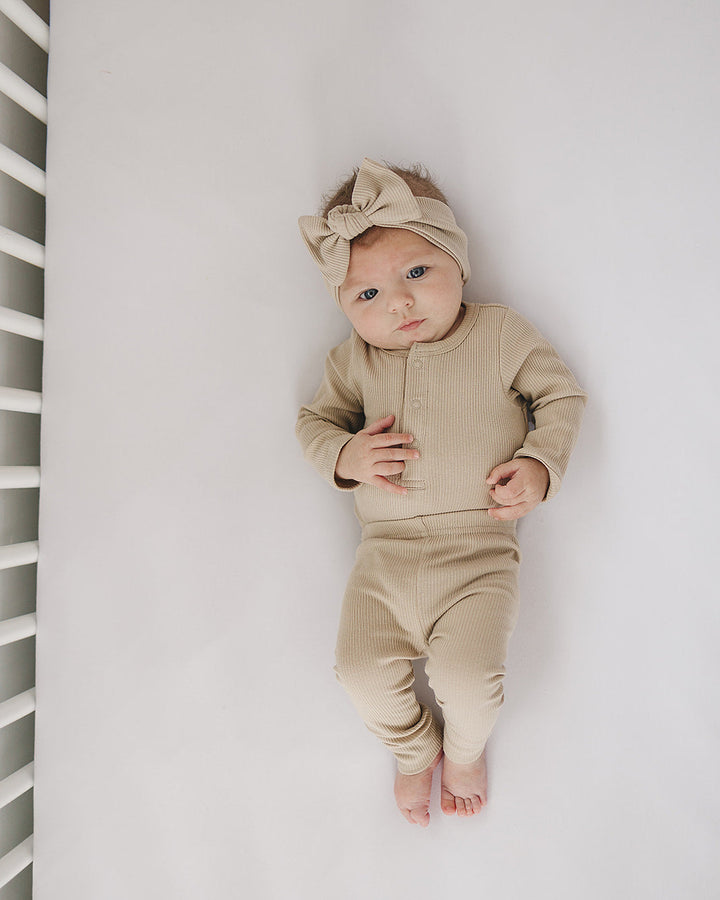 Baby in a beige outfit lying on a white surface with a white crib in the background