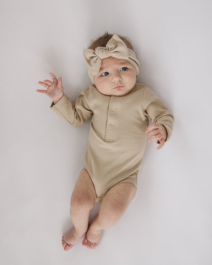 Baby wearing a beige onesie and headband on a light gray background