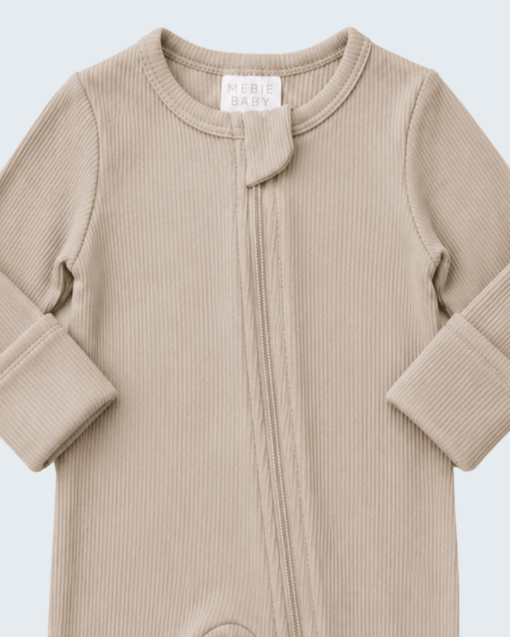 A close-up of a ribbed knit baby footless one-piece sleeper in a light beige color, featuring a front zipper and a small decorative knot at the neckline. The brand label reads &