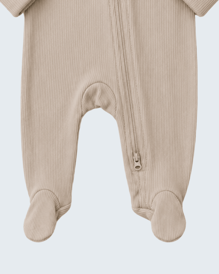 Close-up of a baby footed sleeper in a light beige ribbed fabric, showing the front zipper and foot cuffs against a light gray background.