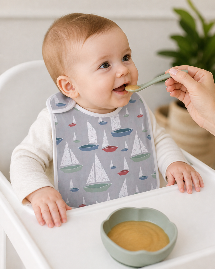 Baby in a high chair with a bib, being fed by an adult hand, with a bowl of food on the tray.