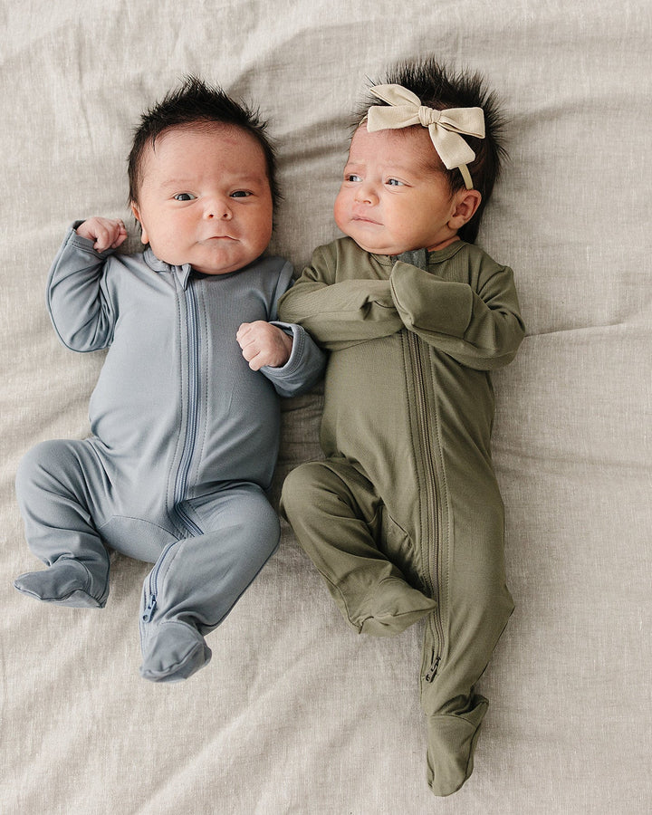 Two babies in gray and green outfits lying on a beige blanket.