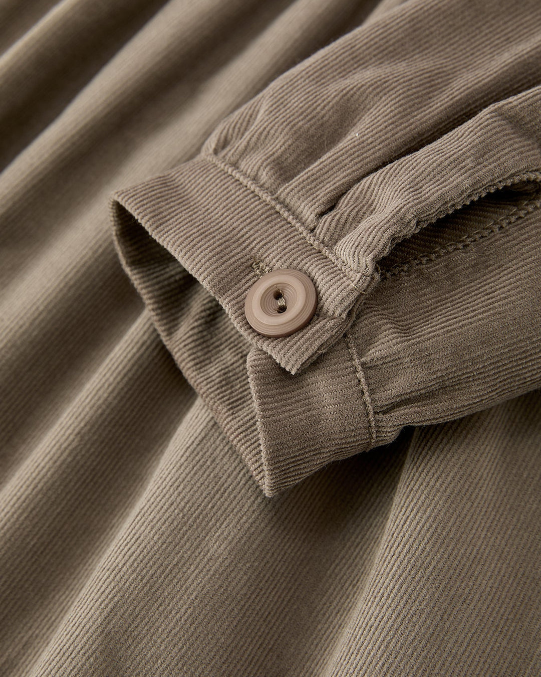 Close-up of a corduroy garment in a warm taupe-beige color, showing the ribbed fabric texture and a matching button on the cuff.