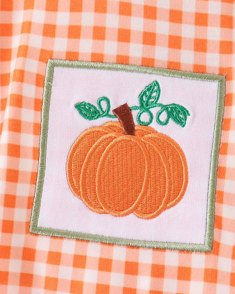 An embroidered pumpkin patch with green leaves and a brown stem on a white square background, placed on an orange and white gingham fabric surface.