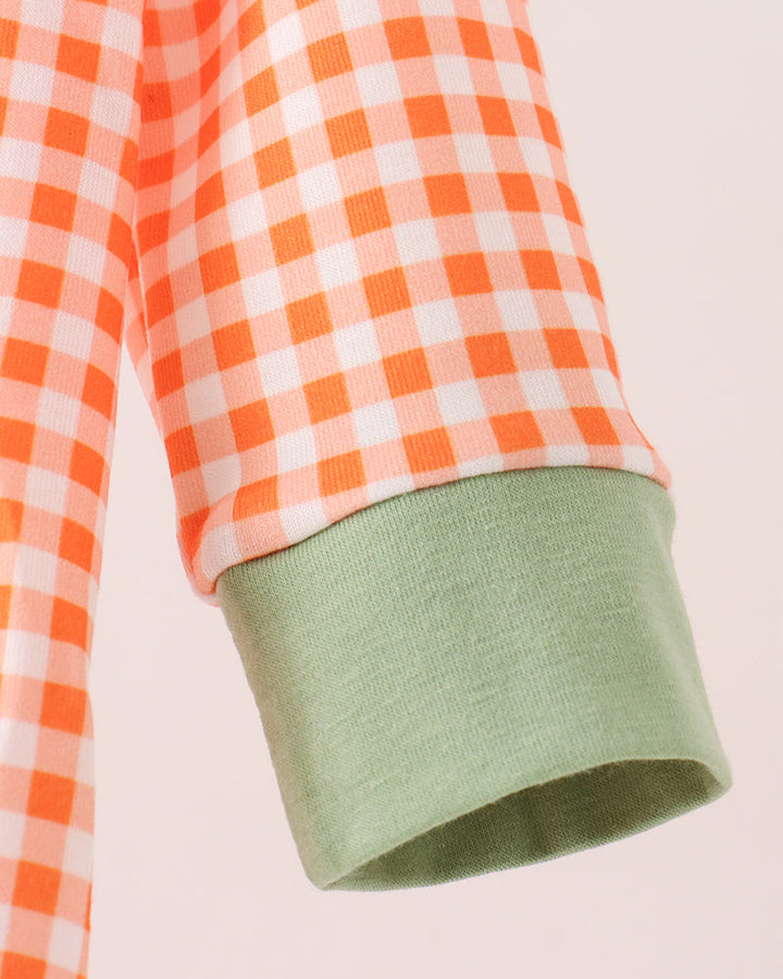 Close-up of a sleeve with an orange and white gingham pattern and a contrasting sage green ribbed cuff, shown against a pale pink background.