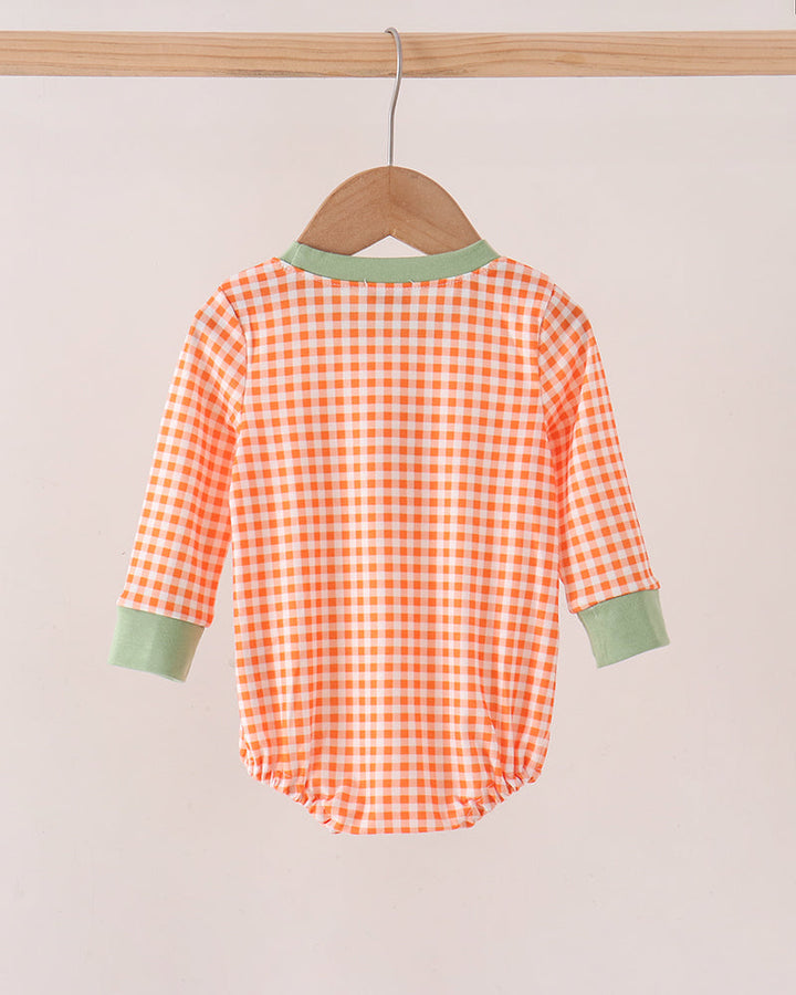 A baby long-sleeve romper with an orange and white gingham check pattern, featuring sage green trim on the collar and cuffs, hanging on a wooden hanger.
