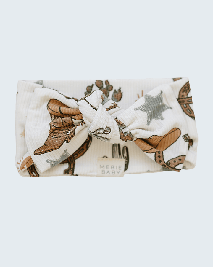 A white muslin baby headband with a large bow, featuring a western-themed print of brown cowboy boots, a gray star, and a horseshoe, with the text &