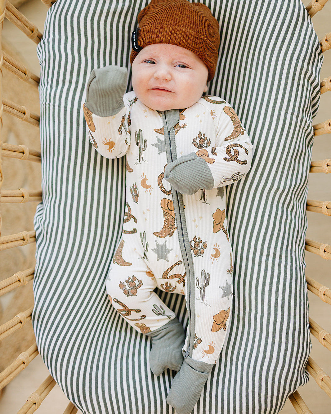 Baby in a striped baby bouncer wearing a cowboy-themed outfit and brown hat.