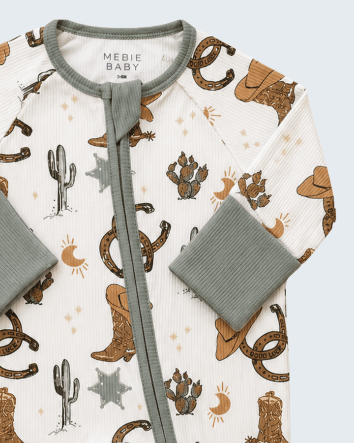 A close-up of a Mebie Baby long-sleeve baby romper featuring a western print with brown cowboy boots, hats, cacti, and snakes on an off-white background, with sage green ribbed trim and cuffs.