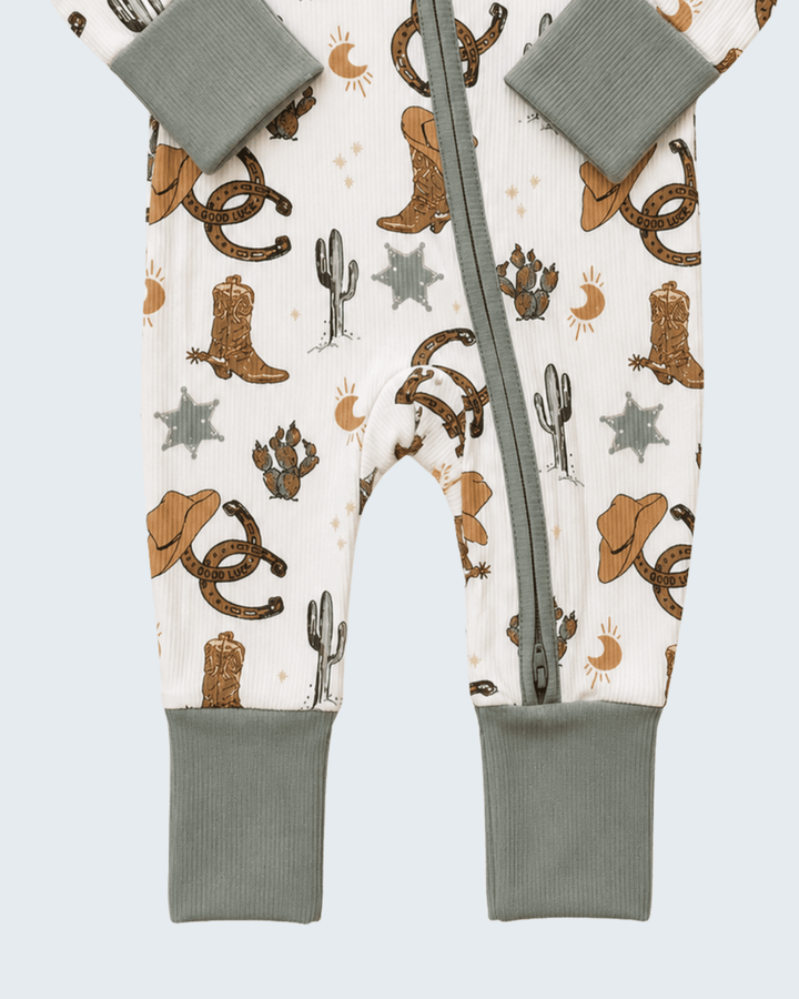 A baby footed sleeper with a western-themed print featuring cowboy boots, hats, horseshoes, cacti, and stars on an off-white background, with sage green ribbed cuffs and a matching zipper.