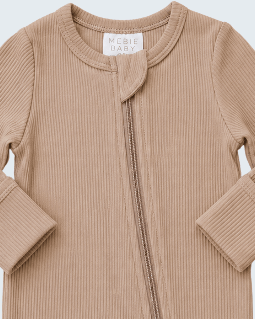 A close-up of a ribbed knit baby zip-up sleeper in a warm beige/tan color, featuring a small decorative tab at the neckline and a white brand label that reads &