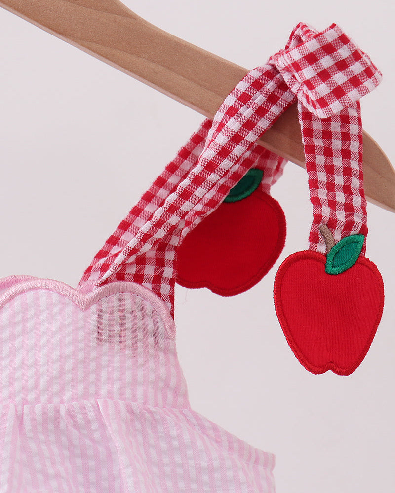 A close-up of a pink and white gingham dress hanging on a wooden hanger, featuring red apple-shaped shoulder straps with green leaf details.