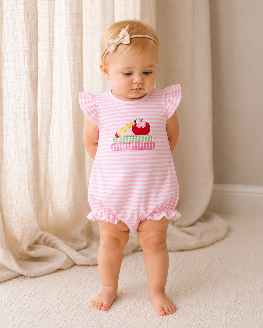 Baby girl is standing in front of linen curtains.