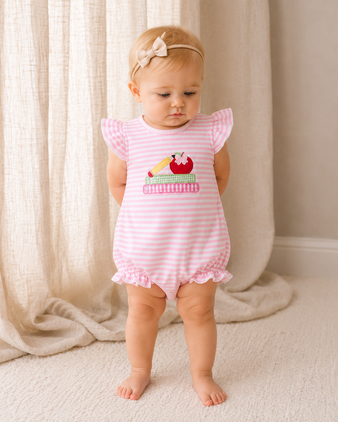 Baby girl is standing in front of linen curtains.