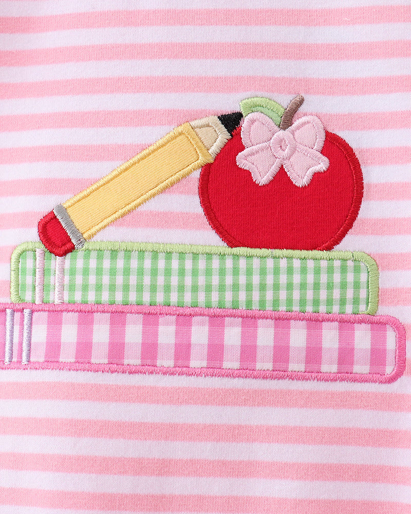 A close-up of a pink and white striped fabric featuring an applique design of a red apple with a pink bow, a yellow pencil, and two books stacked horizontally.