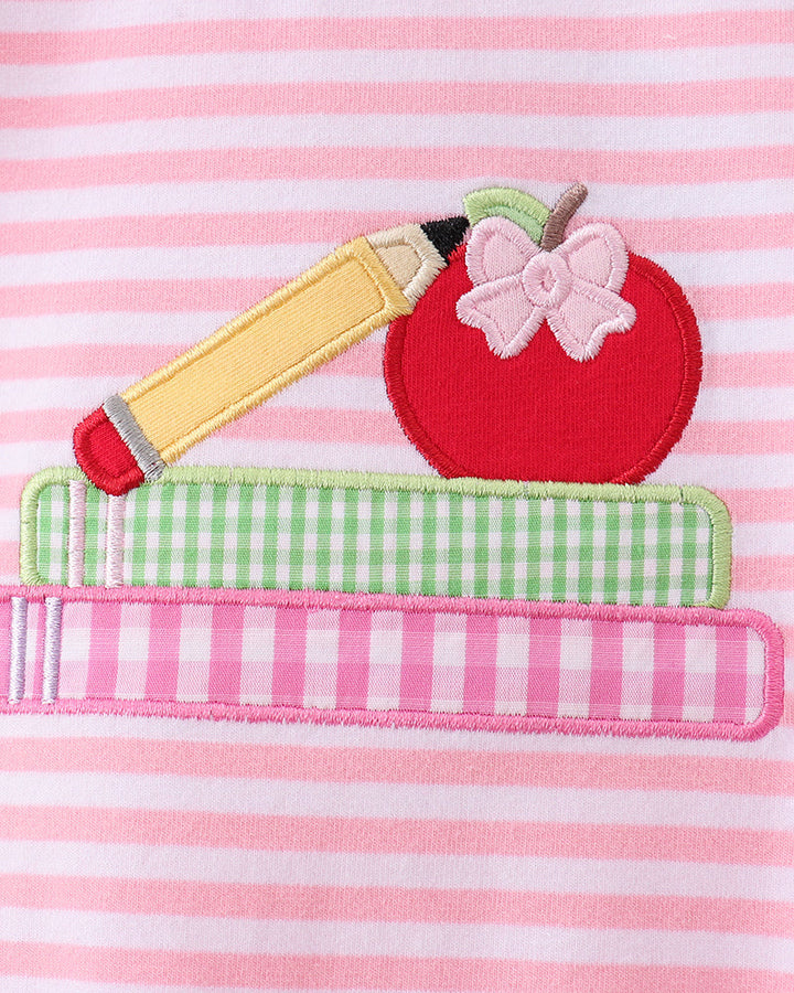 A close-up of a pink and white striped fabric featuring an applique design of a red apple with a pink bow, a yellow pencil, and two books stacked horizontally.