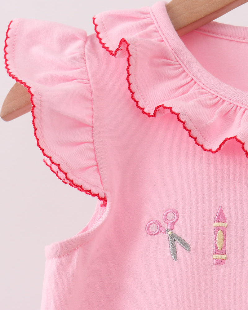 A close-up of a light pink sleeveless top on a wooden hanger, featuring a ruffled collar with red scalloped trim and embroidered scissors and crayon motifs on the front.