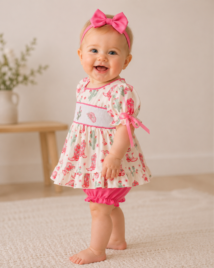 Baby wearing a floral dress with pink accents and a matching headband, standing on a light-colored floor.