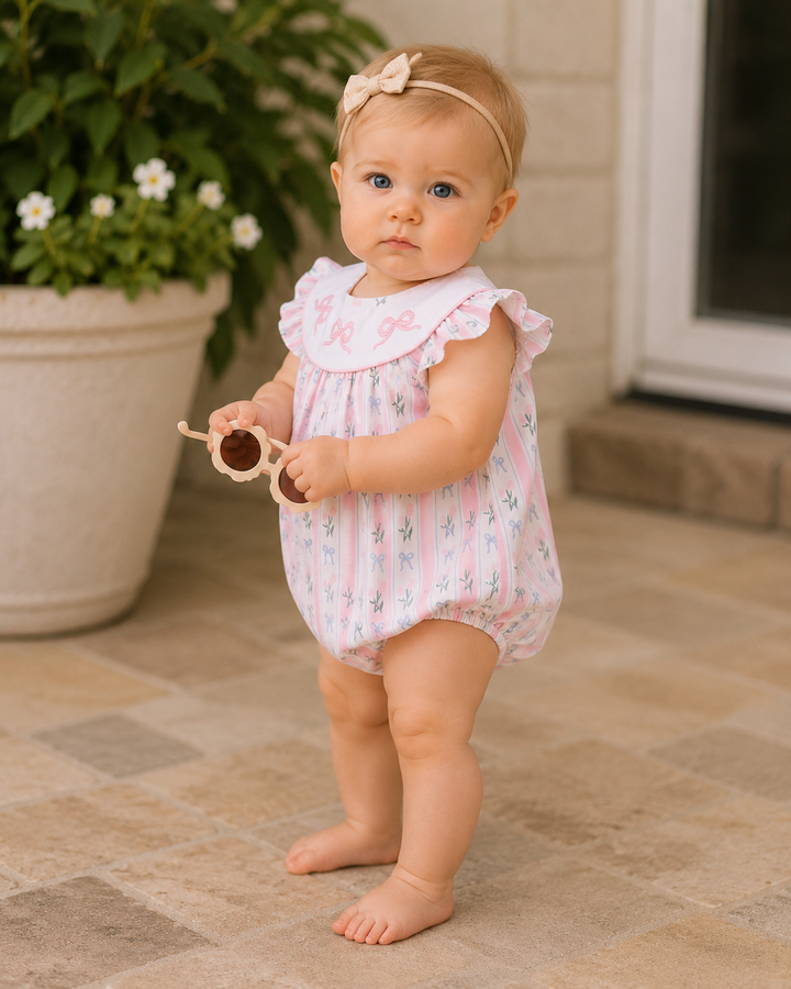Baby in a floral romper standing on a patio with a plant in the background