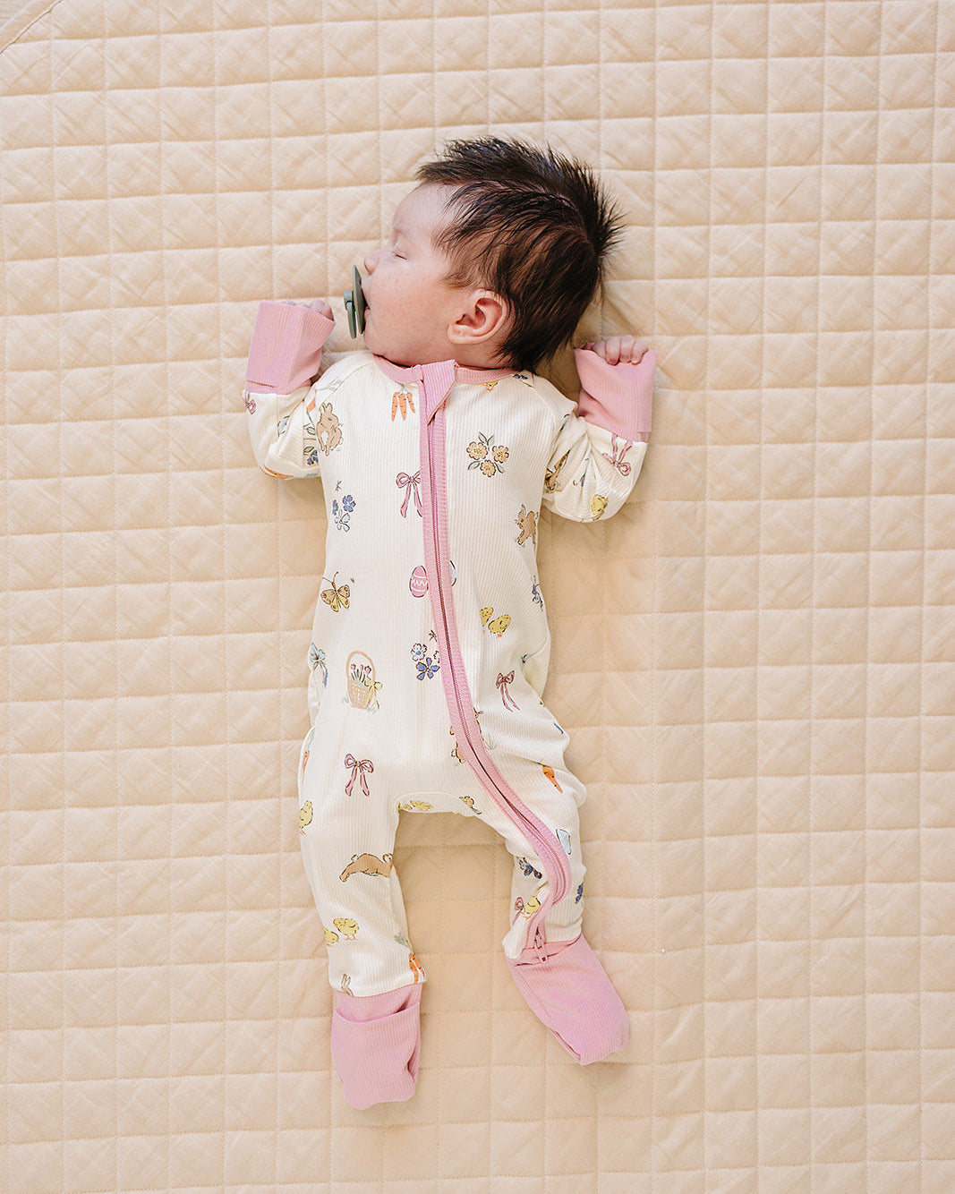 Baby lying on a quilted surface wearing a patterned onesie with pink accents.