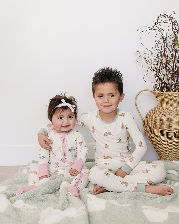 Two children in matching pajamas sitting on a blanket with a decorative vase in the background.