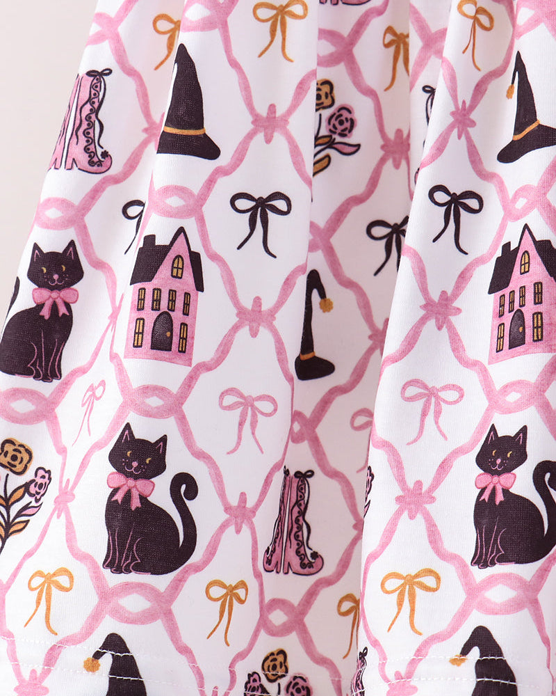 A close-up of a white fabric with a Halloween-themed print featuring black cats, pink houses, pink bows, and gold witch hats.