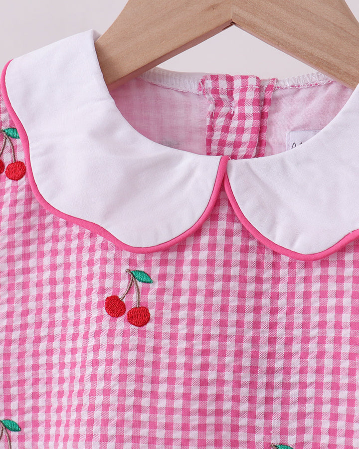 Pink gingham dress with white collar and cherry embroidery on a wooden hanger.