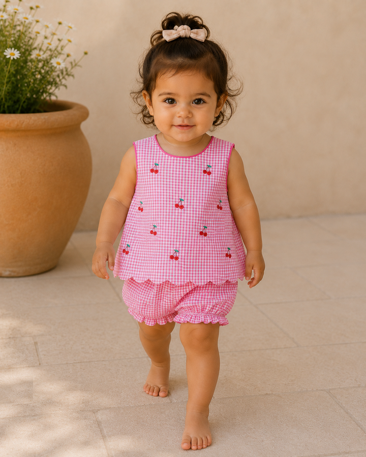 Child wearing a pink checkered outfit with cherry design, standing in front of a potted plant.