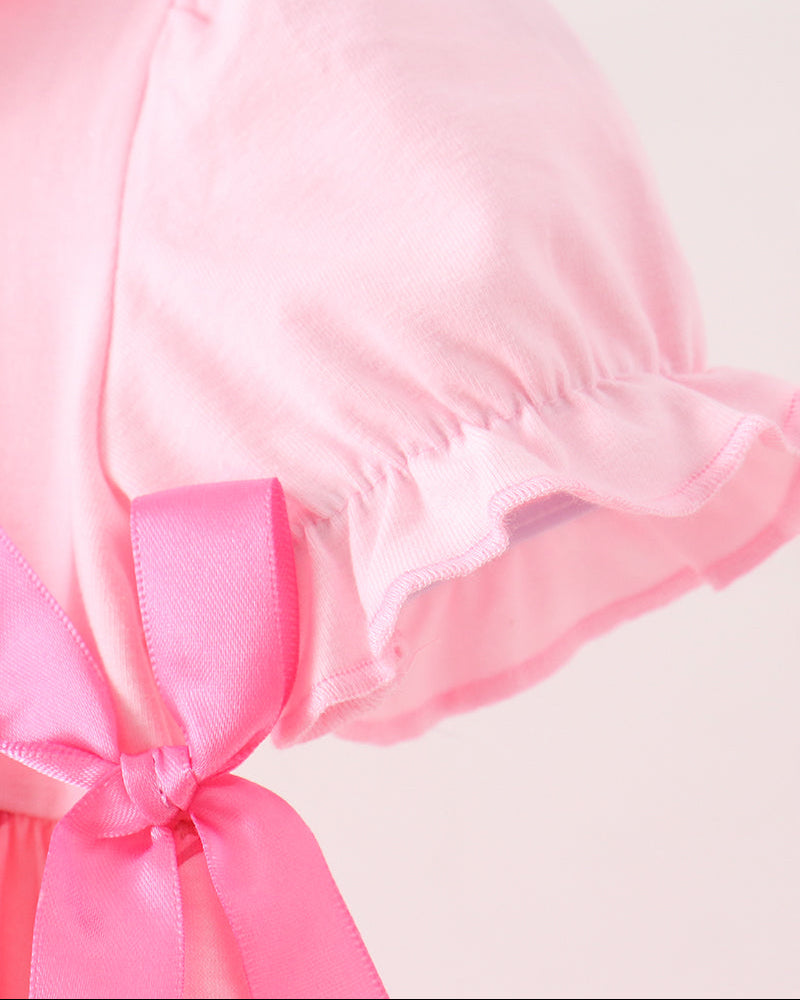 A close-up of a light pink bonnet with ruffled trim and a bright pink satin bow, shown against a pale pink background.