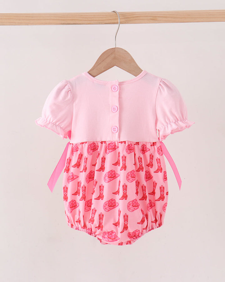 A baby romper hanging on a wooden hanger against a light beige wall. The romper has a light pink short-sleeve top with button closures and a pink patterned bottom featuring cowboy boots and roses.
