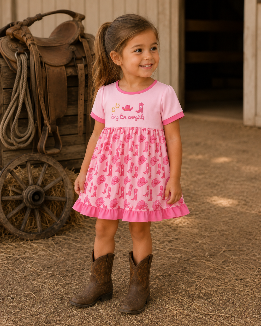 A young girl stands on a straw-covered barn floor wearing a pink short-sleeve dress with a cactus print and cowboy boots, with a rustic wagon visible in the background.
