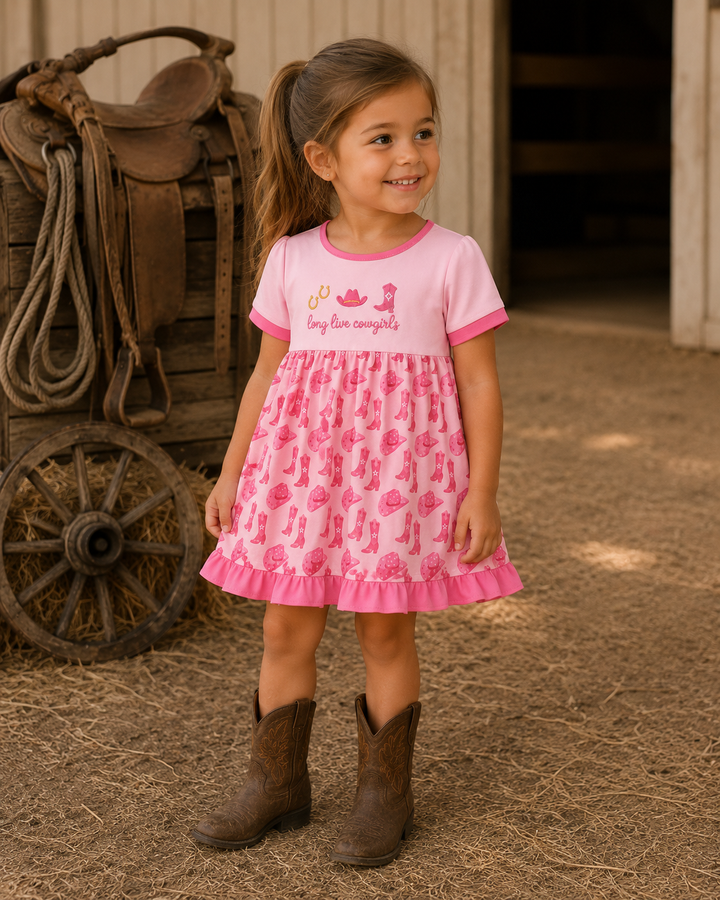 A young girl stands on a straw-covered barn floor wearing a pink short-sleeve dress with a cactus print and cowboy boots, with a rustic wagon visible in the background.