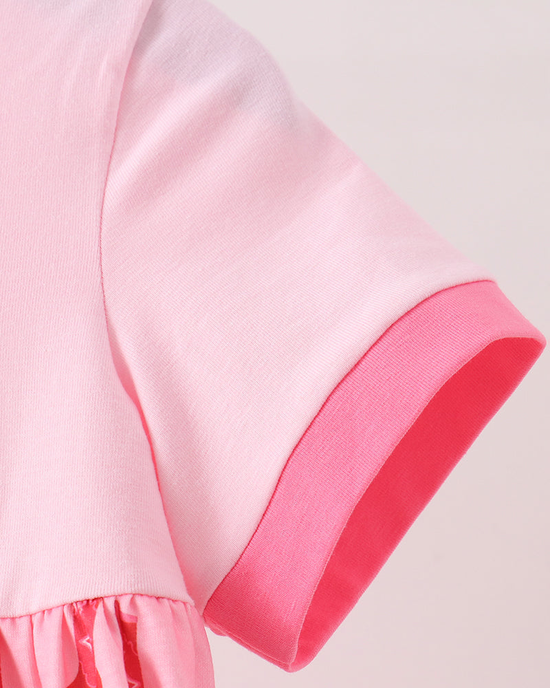 Close-up of a short-sleeve dress or tunic in light pink with a darker pink ribbed cuff and a pink patterned ruffled hem against a pale pink background.