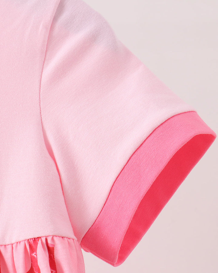 Close-up of a short-sleeve dress or tunic in light pink with a darker pink ribbed cuff and a pink patterned ruffled hem against a pale pink background.