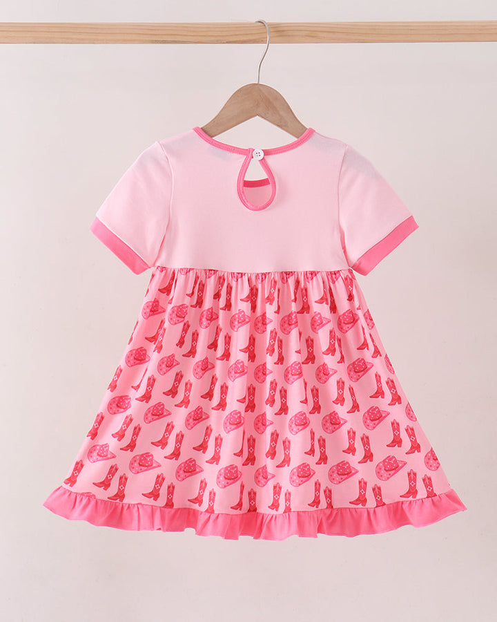 A pink short-sleeve baby dress hanging on a wooden hanger against a light beige wall. The dress has a solid light pink bodice with a small keyhole cutout at the back neck, and a gathered pink skirt printed with a repeating pattern of red cowboy boots and pink roses, finished with a ruffled hem.
