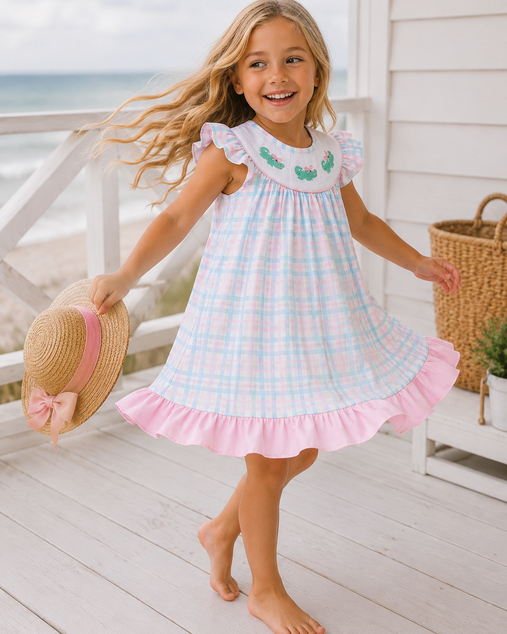 A young girl with long blonde hair stands barefoot on a white wooden porch, wearing a sleeveless pink and light blue plaid dress with a ruffled collar decorated with green embroidered fish. She is holding a straw hat with a pink ribbon.