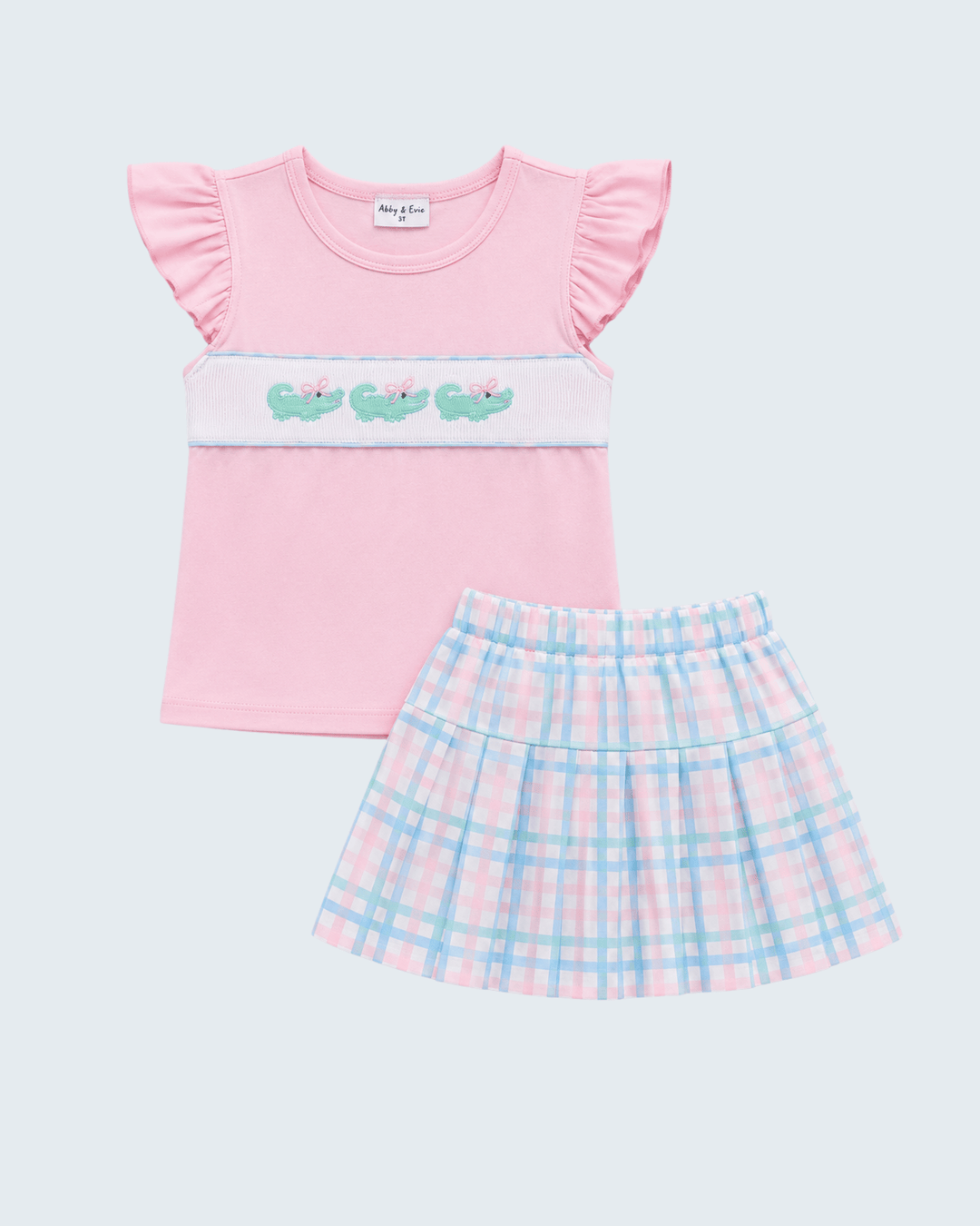 A two-piece girls' outfit set featuring a light pink short-sleeve top with ruffled sleeves and a white horizontal band decorated with three green alligators, paired with a matching pink and light blue plaid pleated skirt, shown on a light gray background.