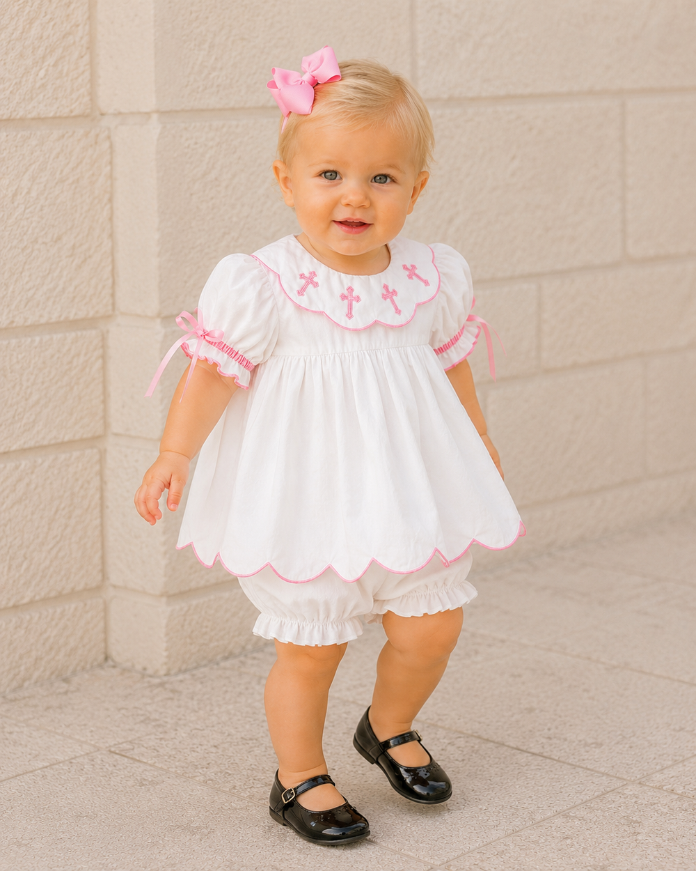 A toddler girl wearing a white baby dress with pink trim, matching white ruffled bloomers, and black Mary Jane shoes, standing against a light beige stone wall.