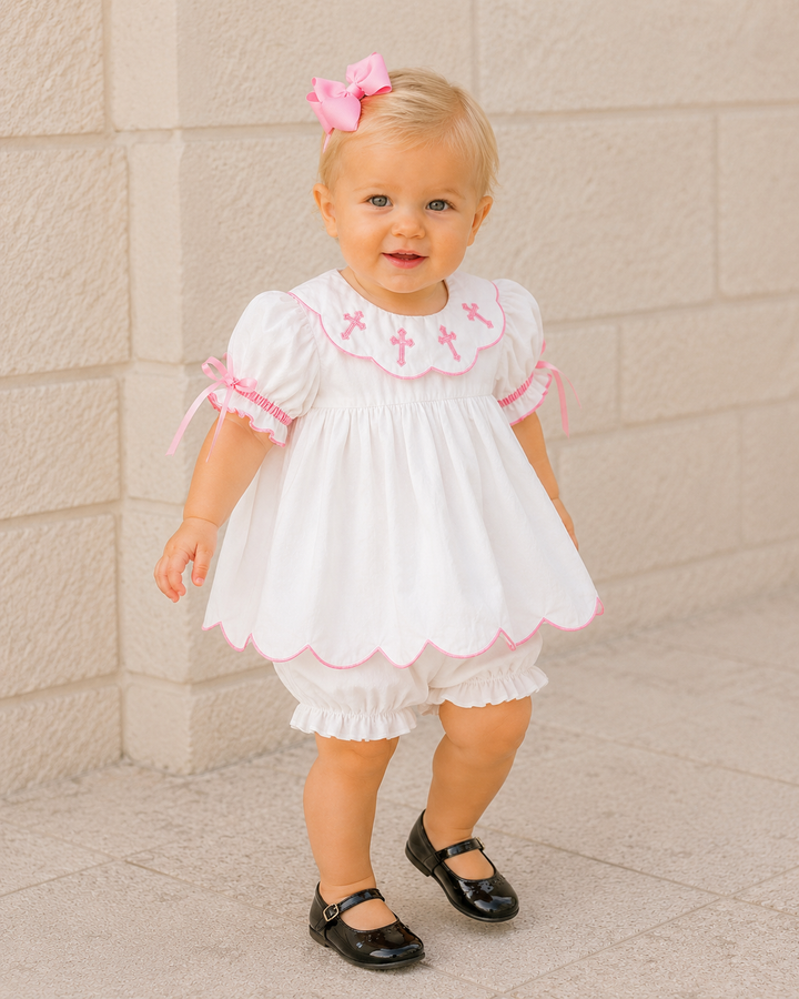 A toddler girl wearing a white baby dress with pink trim, matching white ruffled bloomers, and black Mary Jane shoes, standing against a light beige stone wall.