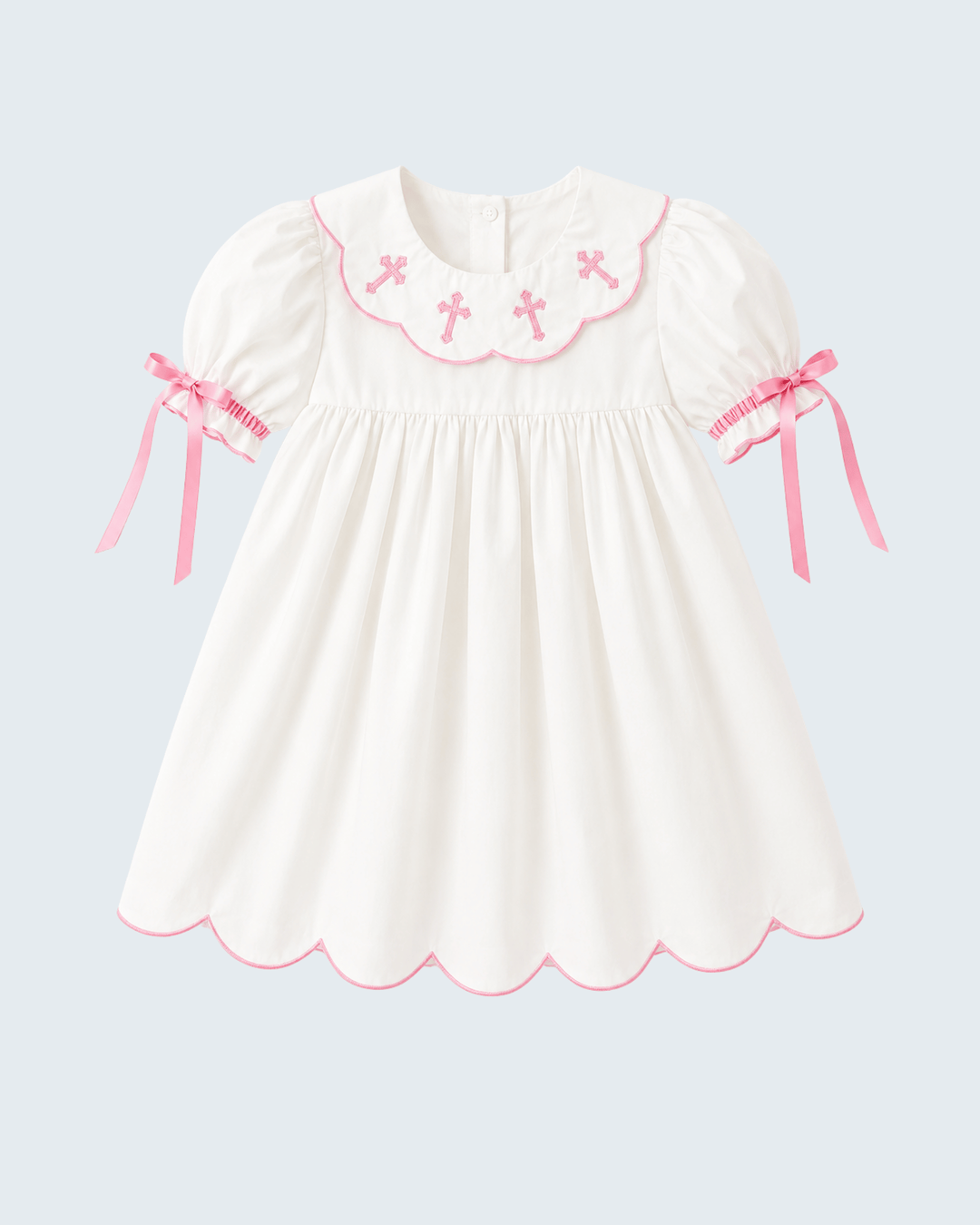 A white short-sleeve baby dress with pink trim, featuring a scalloped Peter Pan collar decorated with pink embroidered crosses, pink ribbon bows on the sleeves, and a scalloped hem, shown against a light gray background.