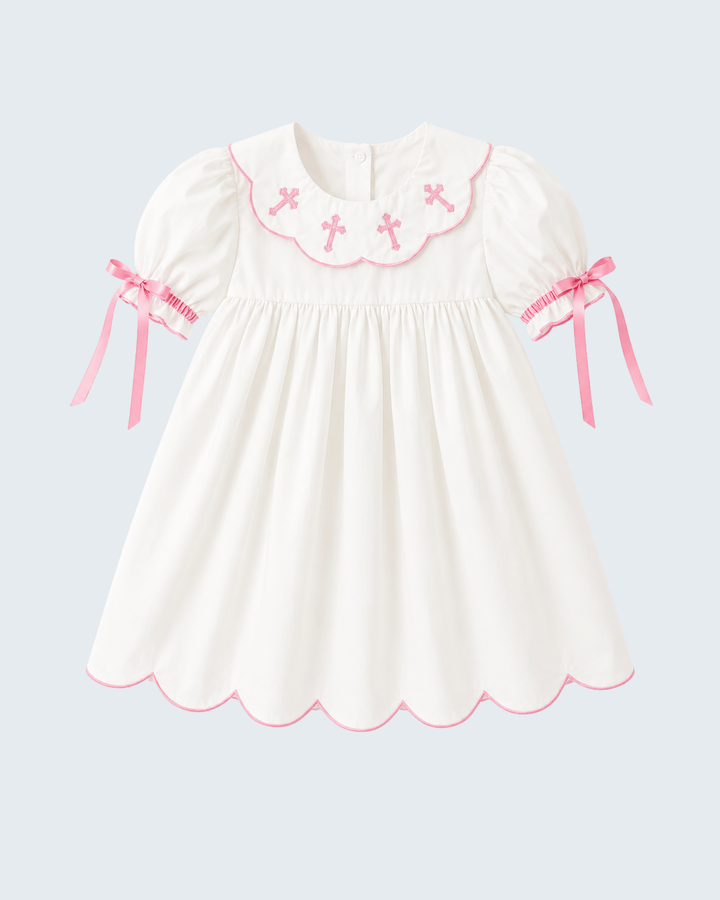 A white short-sleeve baby dress with pink trim, featuring a scalloped Peter Pan collar decorated with pink embroidered crosses, pink ribbon bows on the sleeves, and a scalloped hem, shown against a light gray background.