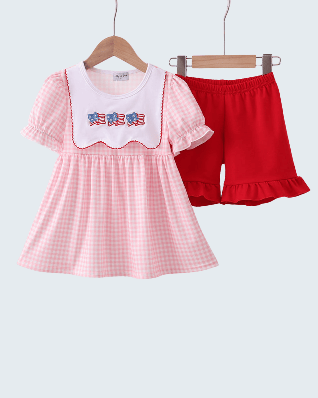 Pink checkered dress with white collar and red shorts on hangers against a light gray background