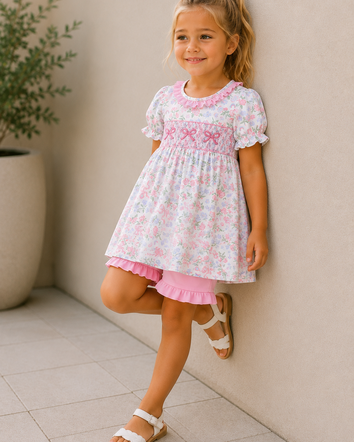 Young girl in a floral dress standing against a beige wall with a plant in the background.