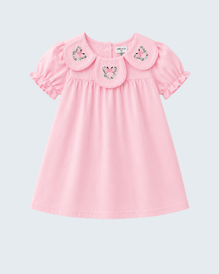 A light pink short-sleeve baby dress with a large Peter Pan collar decorated with three embroidered floral motifs, shown against a light gray background.