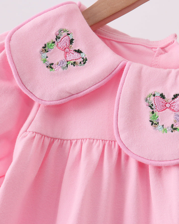 A close-up of a pink long-sleeve dress with a large rounded Peter Pan collar, featuring embroidered floral bow motifs on both collar points, hanging on a wooden hanger against a light background.