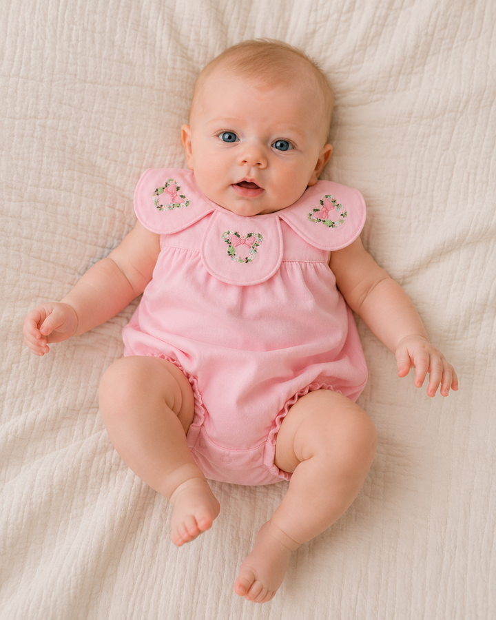 A baby lying on a textured cream-colored blanket, wearing a light pink sleeveless romper with ruffled collar details and embroidered floral motifs.